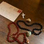 Two beaded necklaces on a wooden surface with an envelope and small gold object.