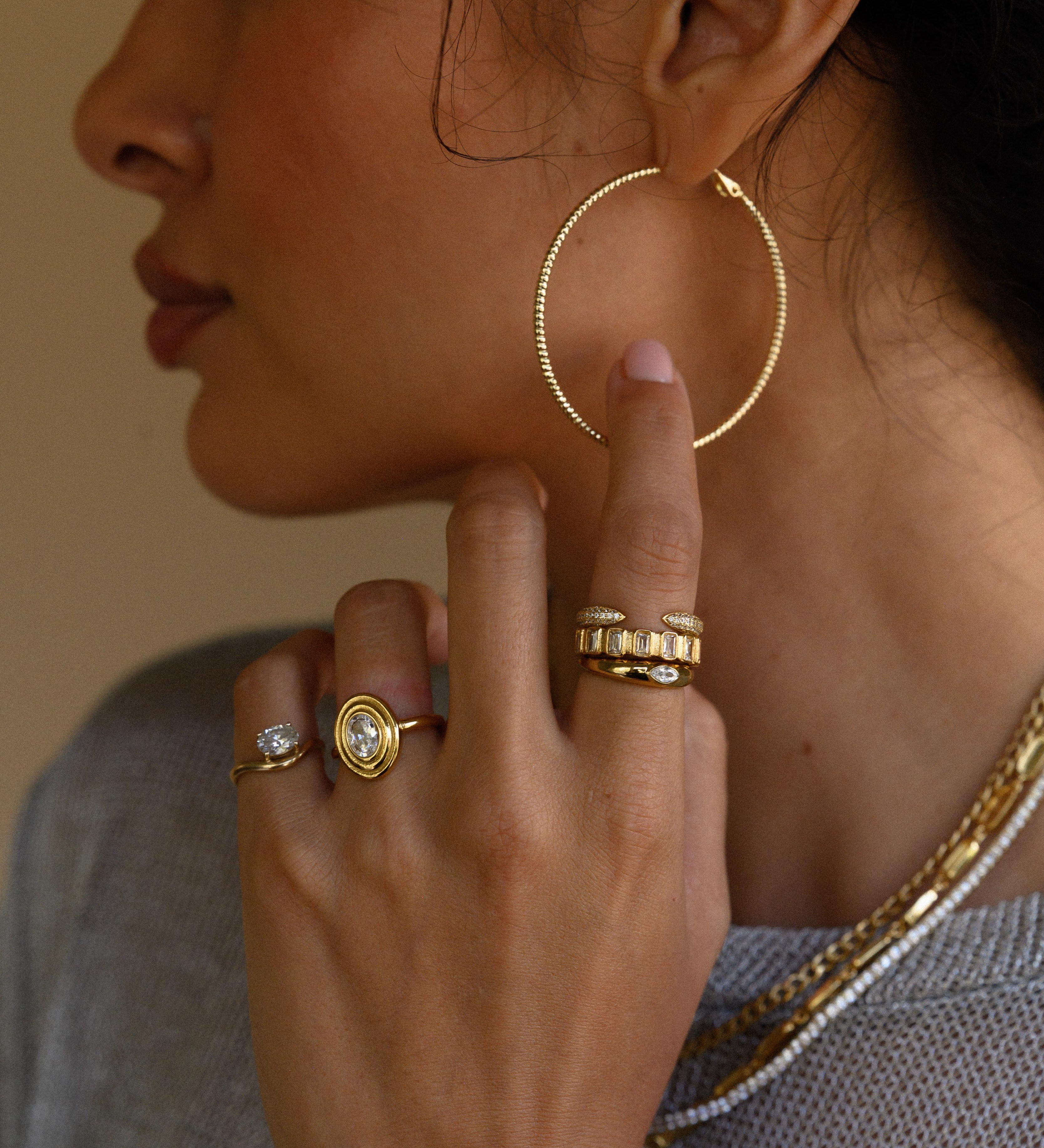 Close-up side view of a woman wearing gold hoops, rings, and necklaces in a grey sweater 