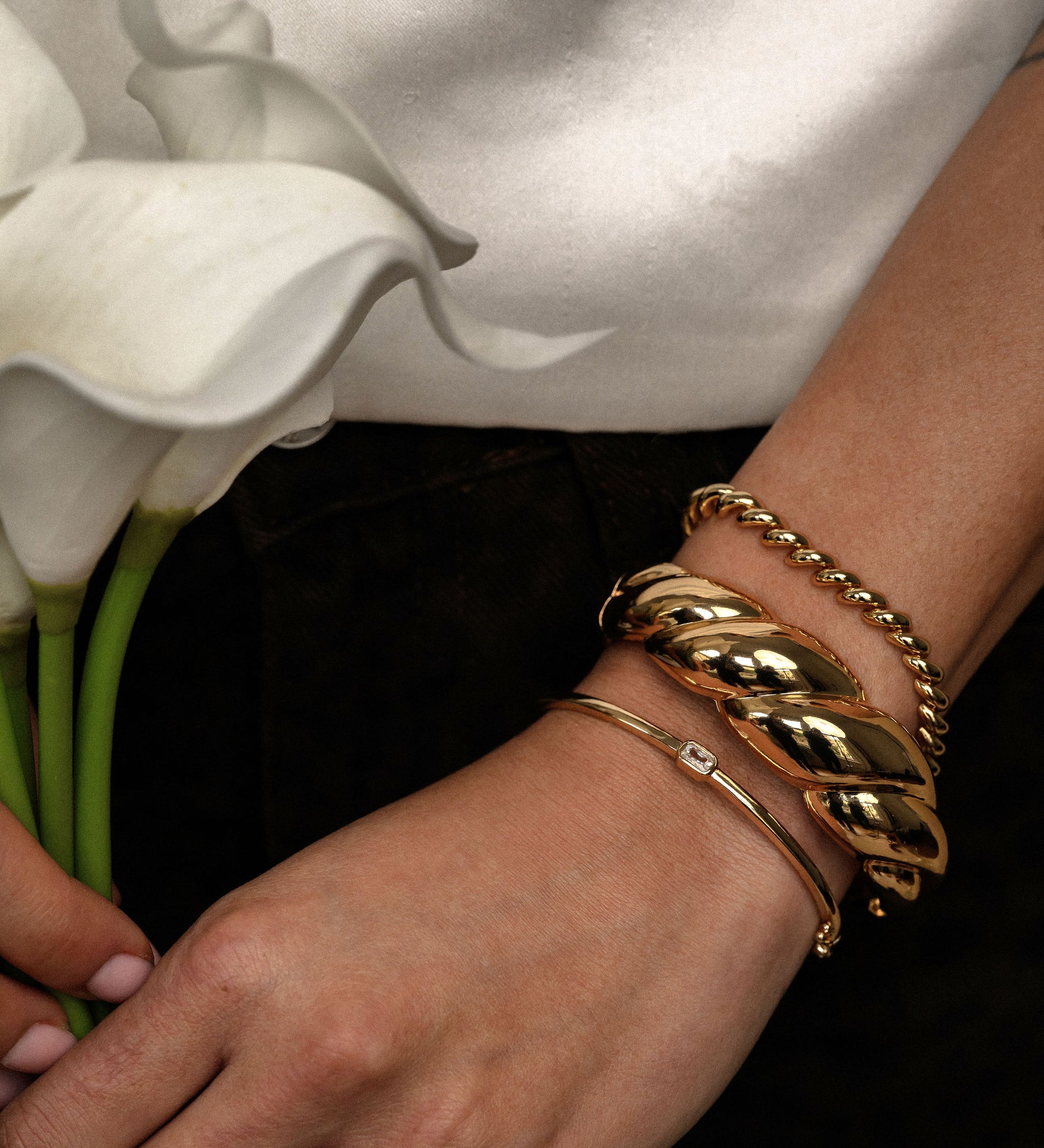 Gold bracelets on a wrist with a background of white flowers and a blurred white shirt.