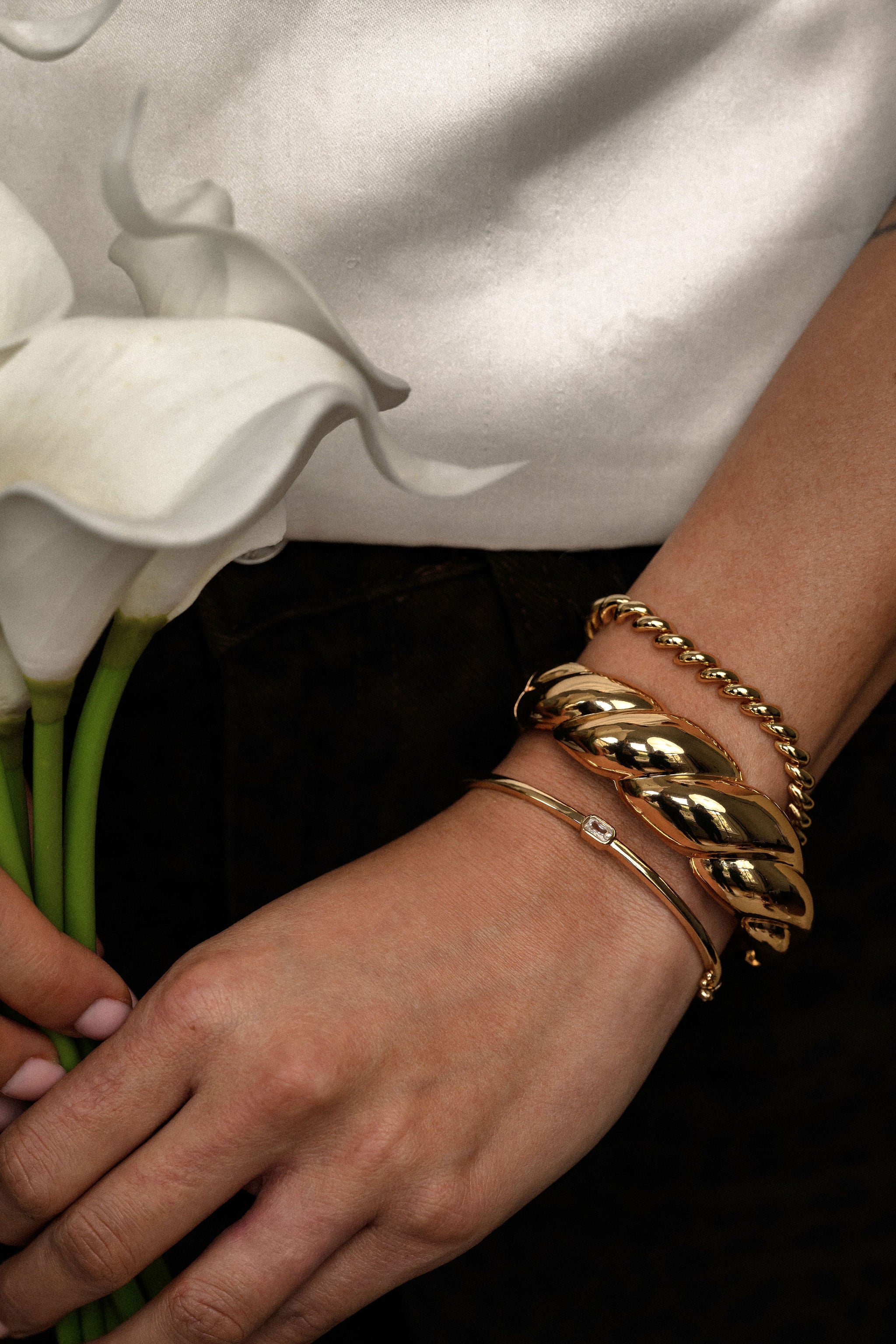 Gold bracelets on a wrist with a background of white flowers and a blurred white shirt.