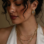 Woman wearing gold jewelry including earrings and necklaces against a neutral background