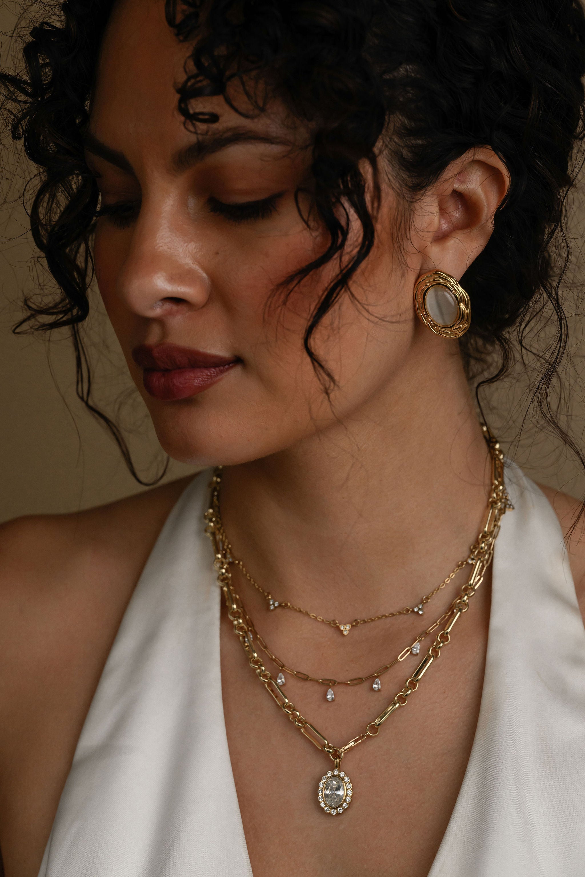 Woman wearing gold jewelry including earrings and necklaces against a neutral background