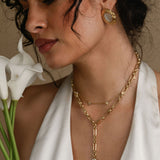 Woman wearing gold necklaces and earrings, holding white flowers, against a neutral background