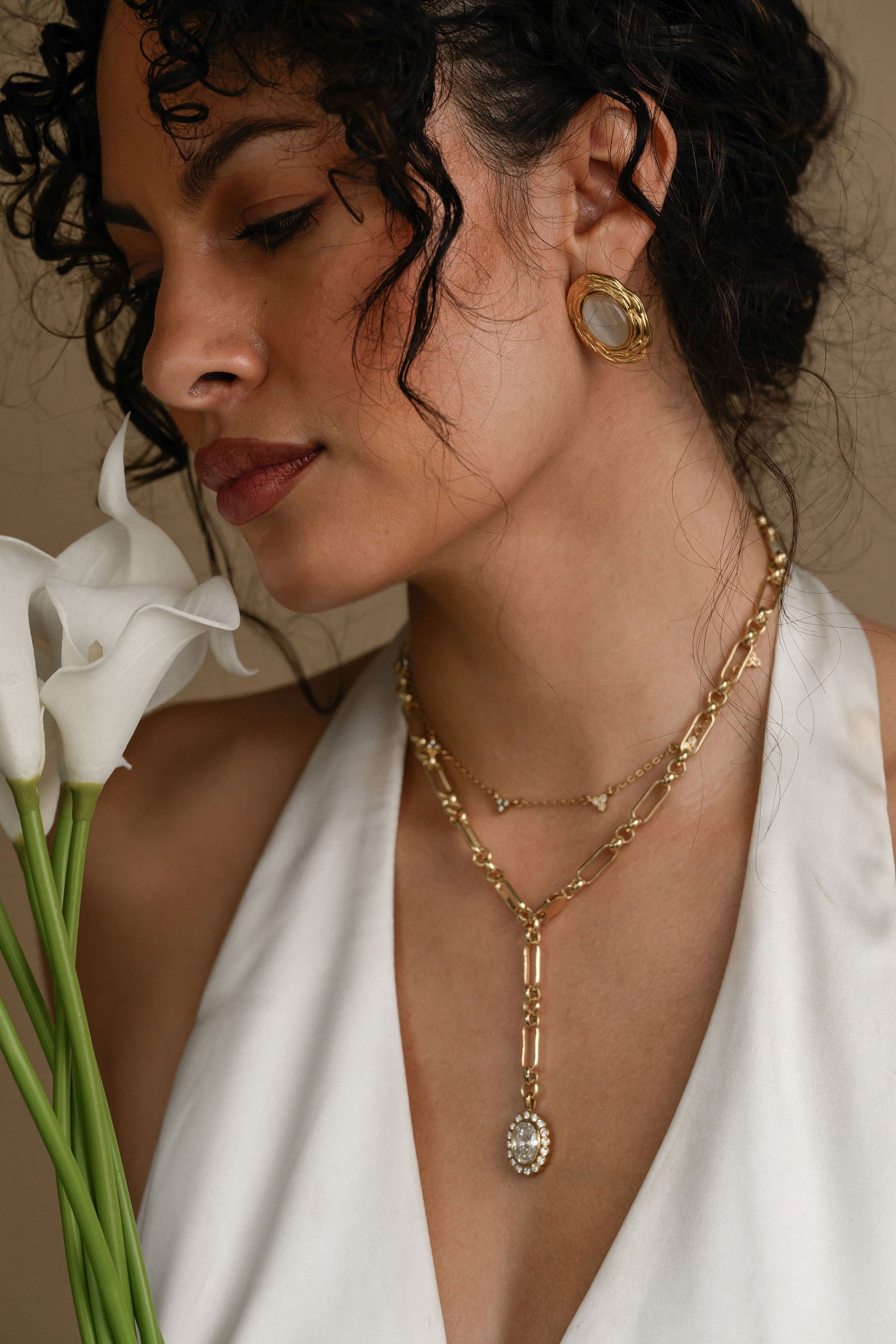 Woman wearing gold necklaces and earrings, holding white flowers, against a neutral background