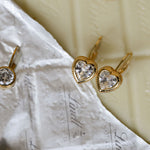 Gold heart-shaped earrings with clear gemstones on a textured white surface.