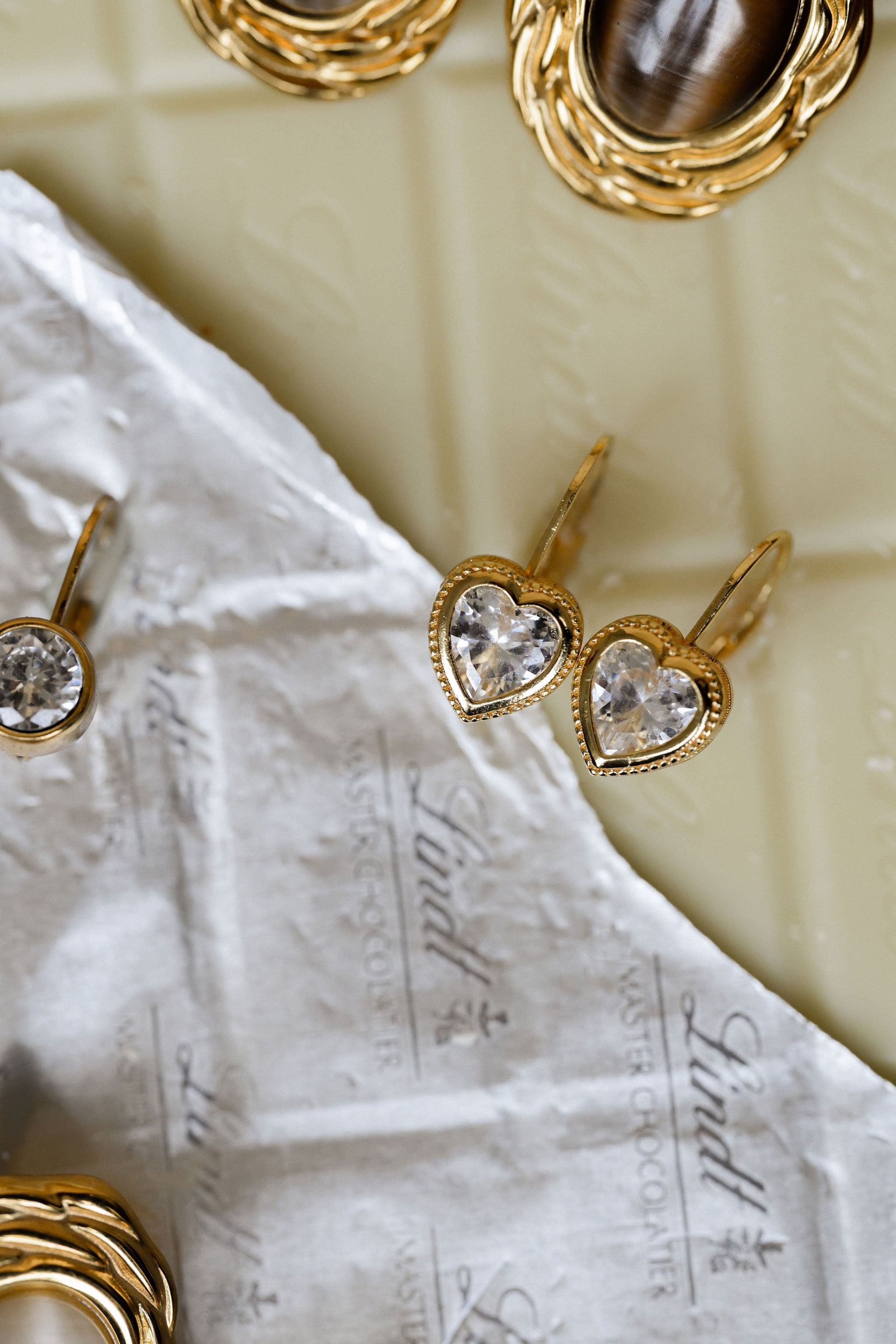 Gold heart-shaped earrings with clear gemstones on a textured white surface.