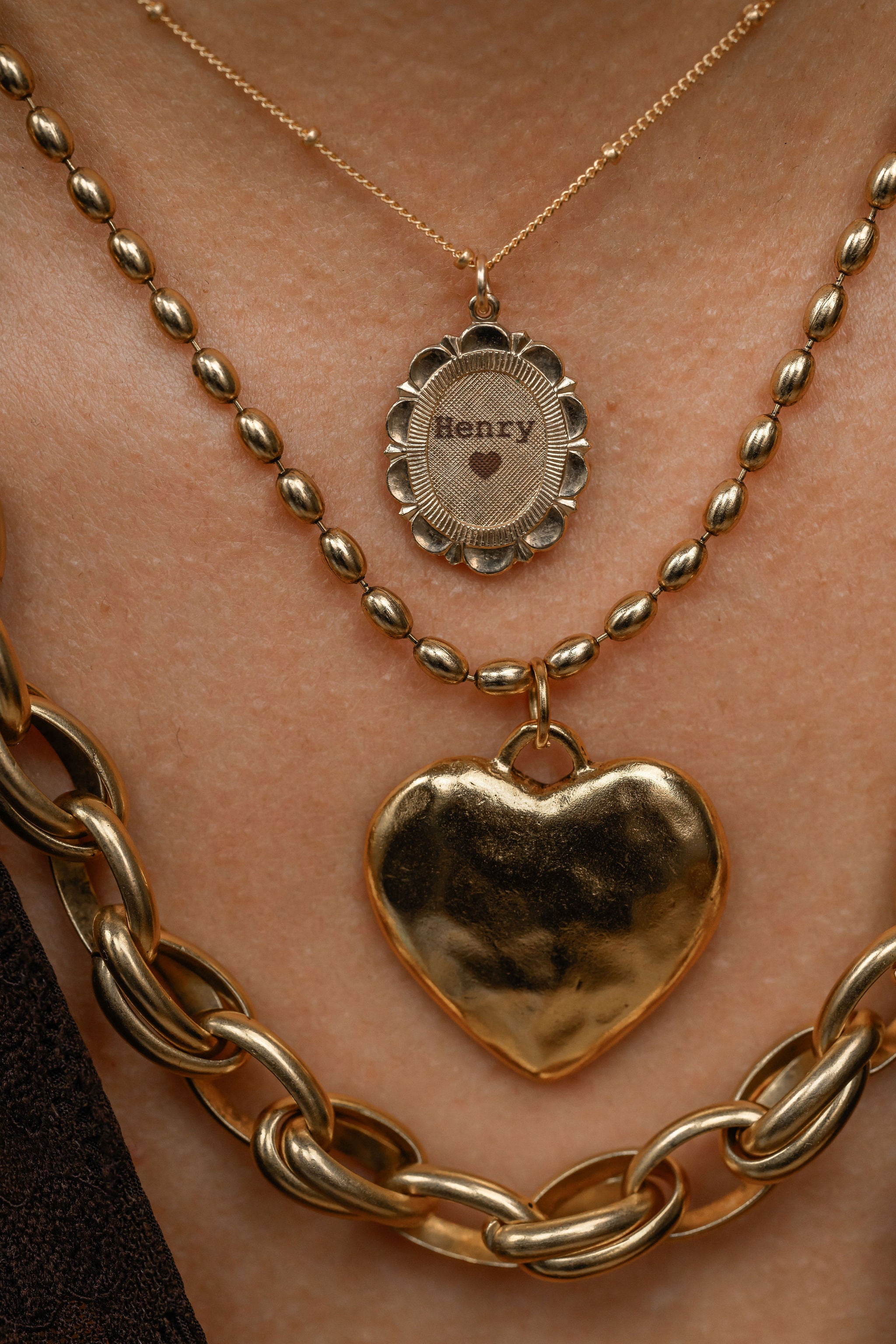Gold necklaces with heart pendant and 'Henry' pendant on a person's neck.