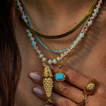 Close-up of a person wearing layered necklaces, including a gold fish pendant, a turquoise stone pendant, and beaded strands in turquoise, cream, and pink, along with gold rings.
