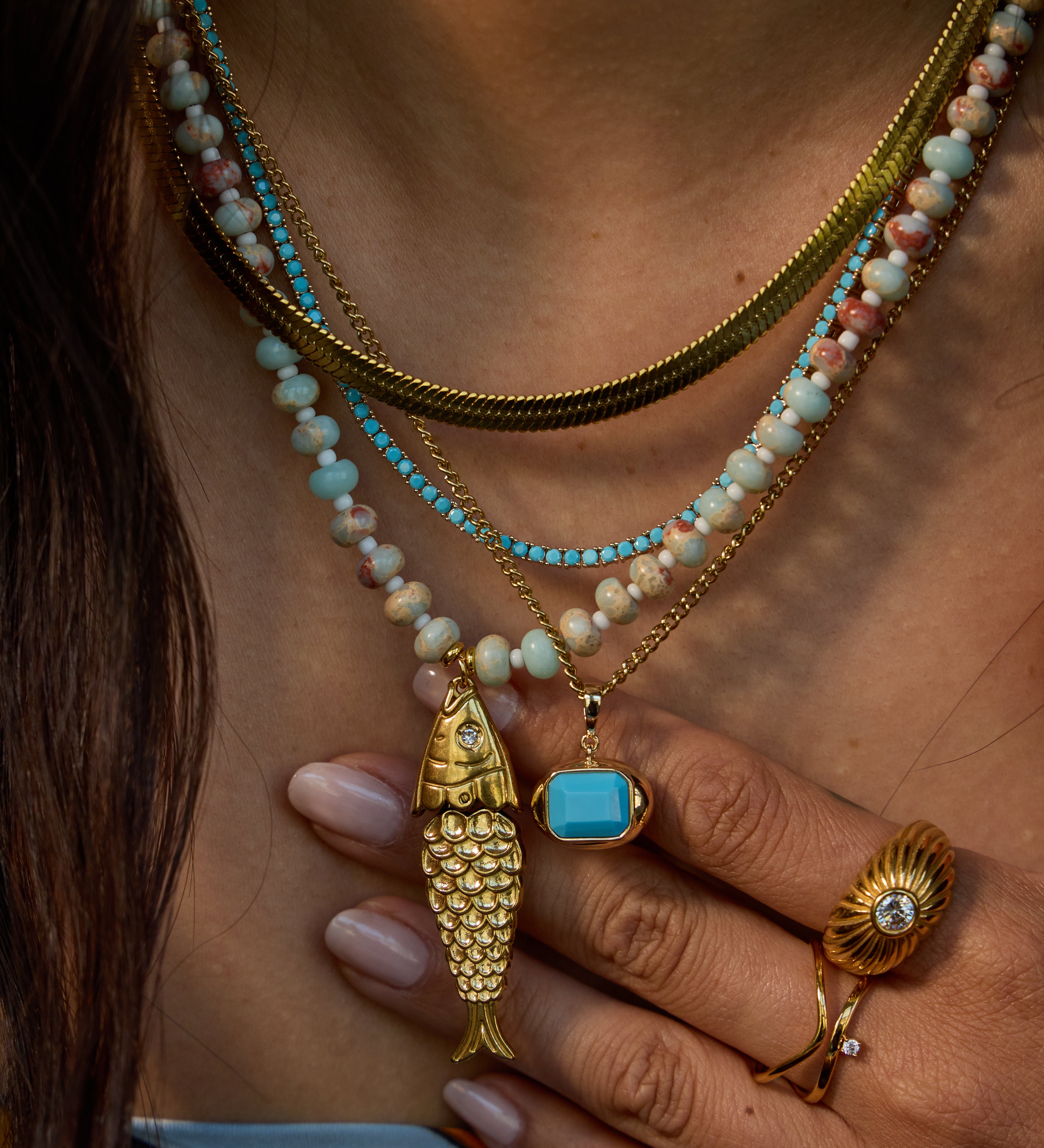 Close-up of a person wearing layered necklaces, including a gold fish pendant, a turquoise stone pendant, and beaded strands in turquoise, cream, and pink, along with gold rings.