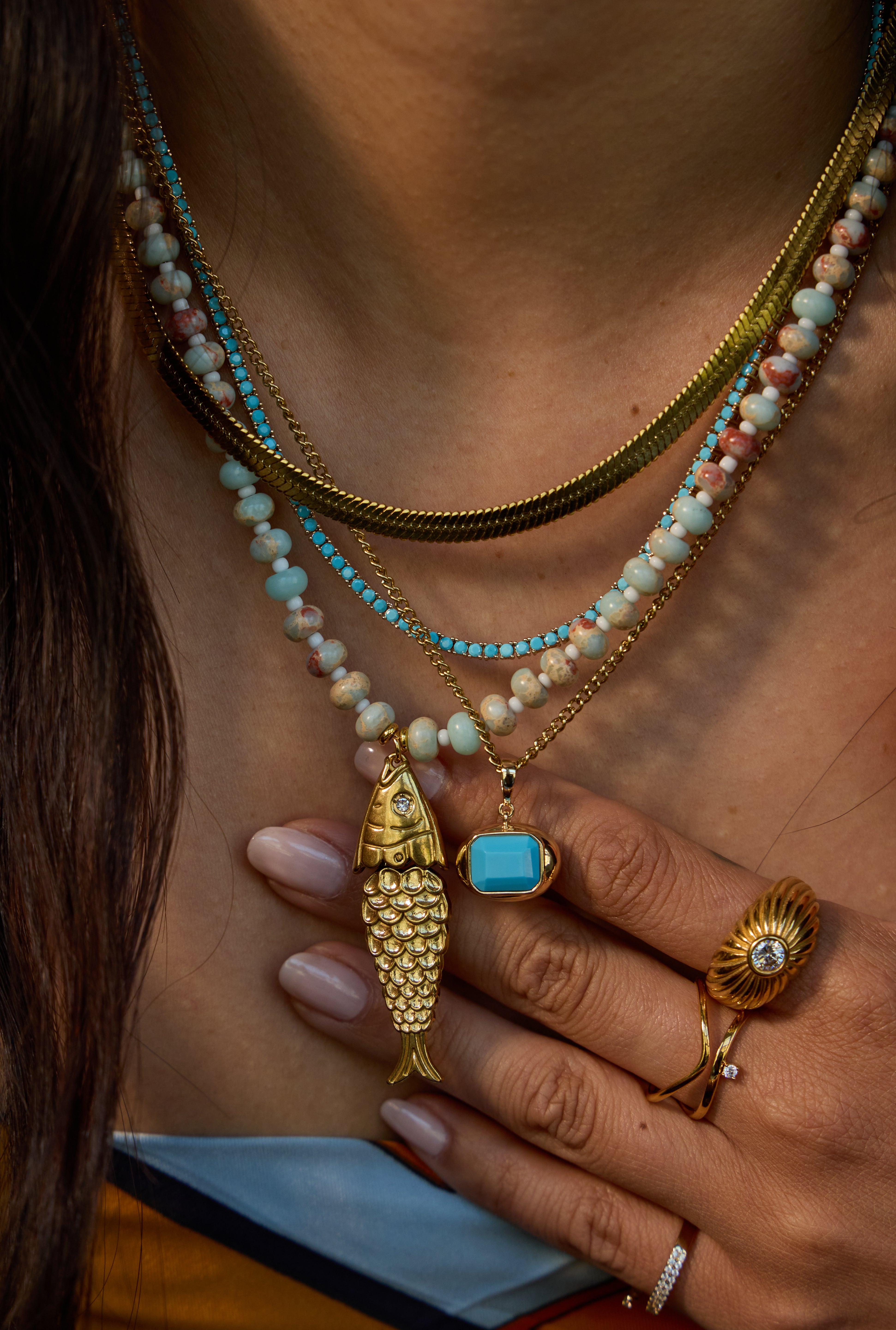 Close-up of a person wearing layered necklaces, including a gold fish pendant, a turquoise stone pendant, and beaded strands in turquoise, cream, and pink, along with gold rings.