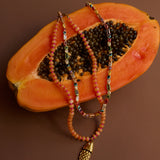 Two beaded necklaces with a gold fish charm on a halved papaya against a brown background