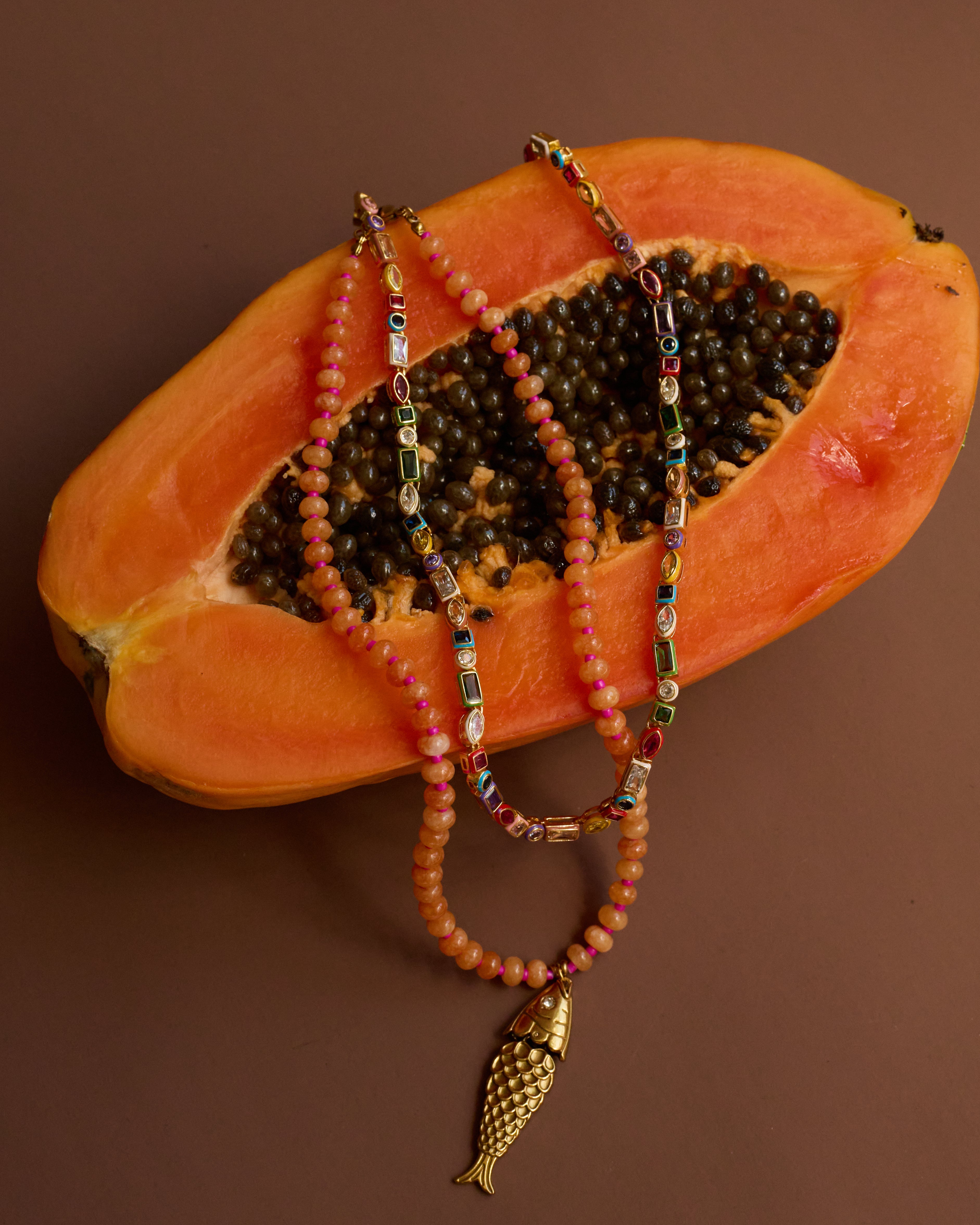 Two beaded necklaces with a gold fish charm on a halved papaya against a brown background