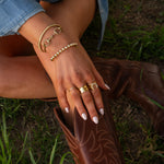 Close-up of a hand with multiple gold bracelets and rings on a brown leather boot against a grassy background.