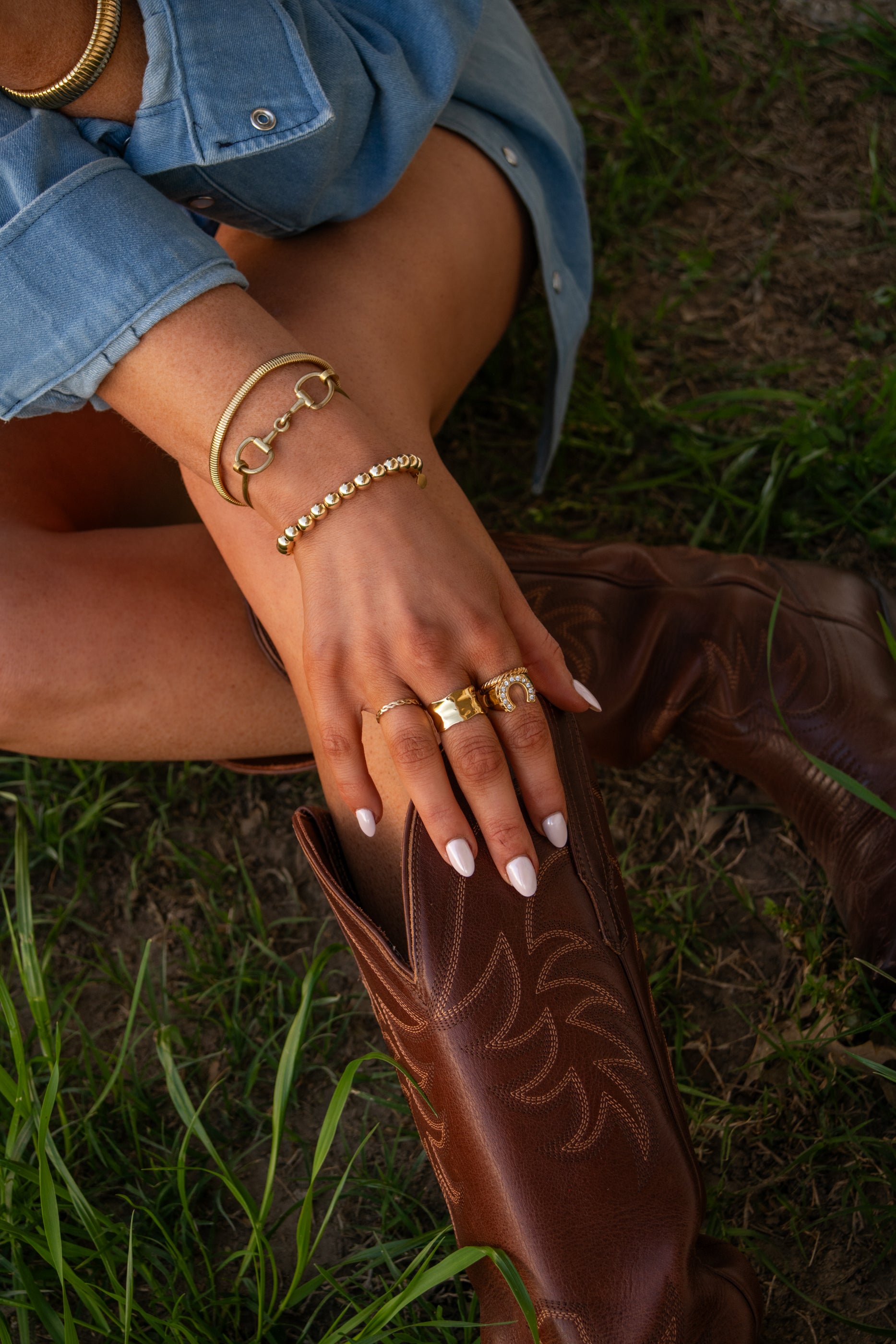 Close-up of a hand with multiple gold bracelets and rings on a brown leather boot against a grassy background.