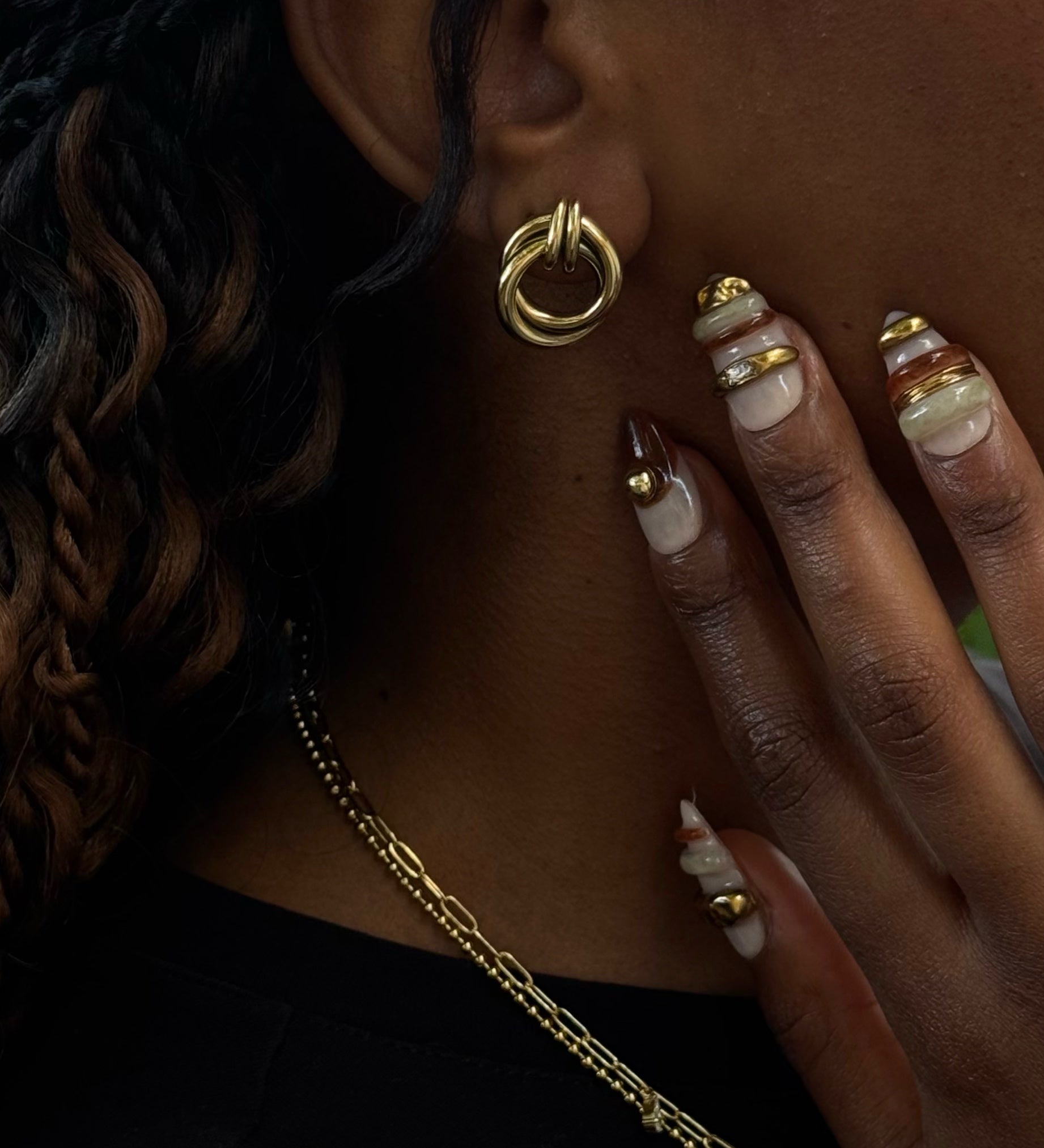 Side shot of a woman with curly hair wearing statement earring, with accented metallic nails and gold necklaces 