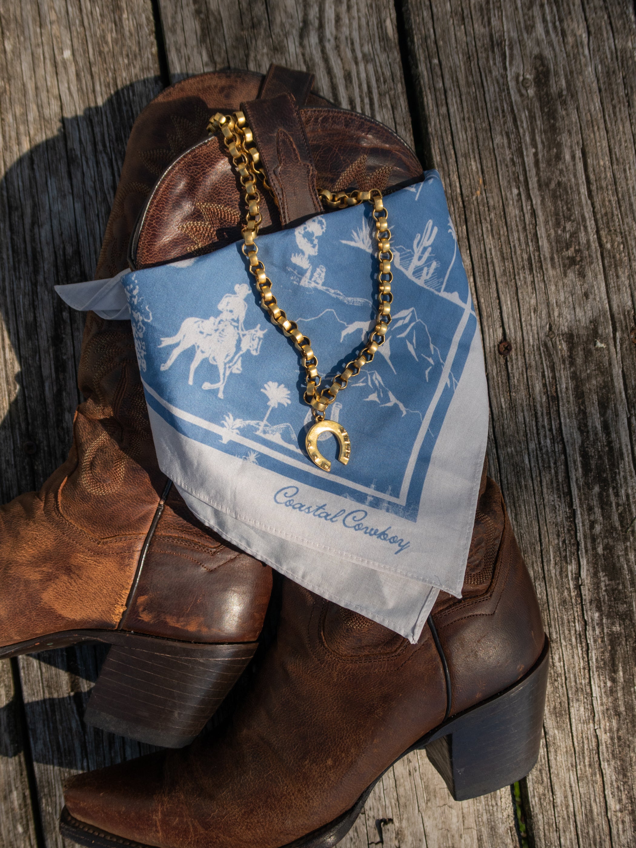 Brown cowboy boots with a blue bandana and gold necklace on a wooden surface
