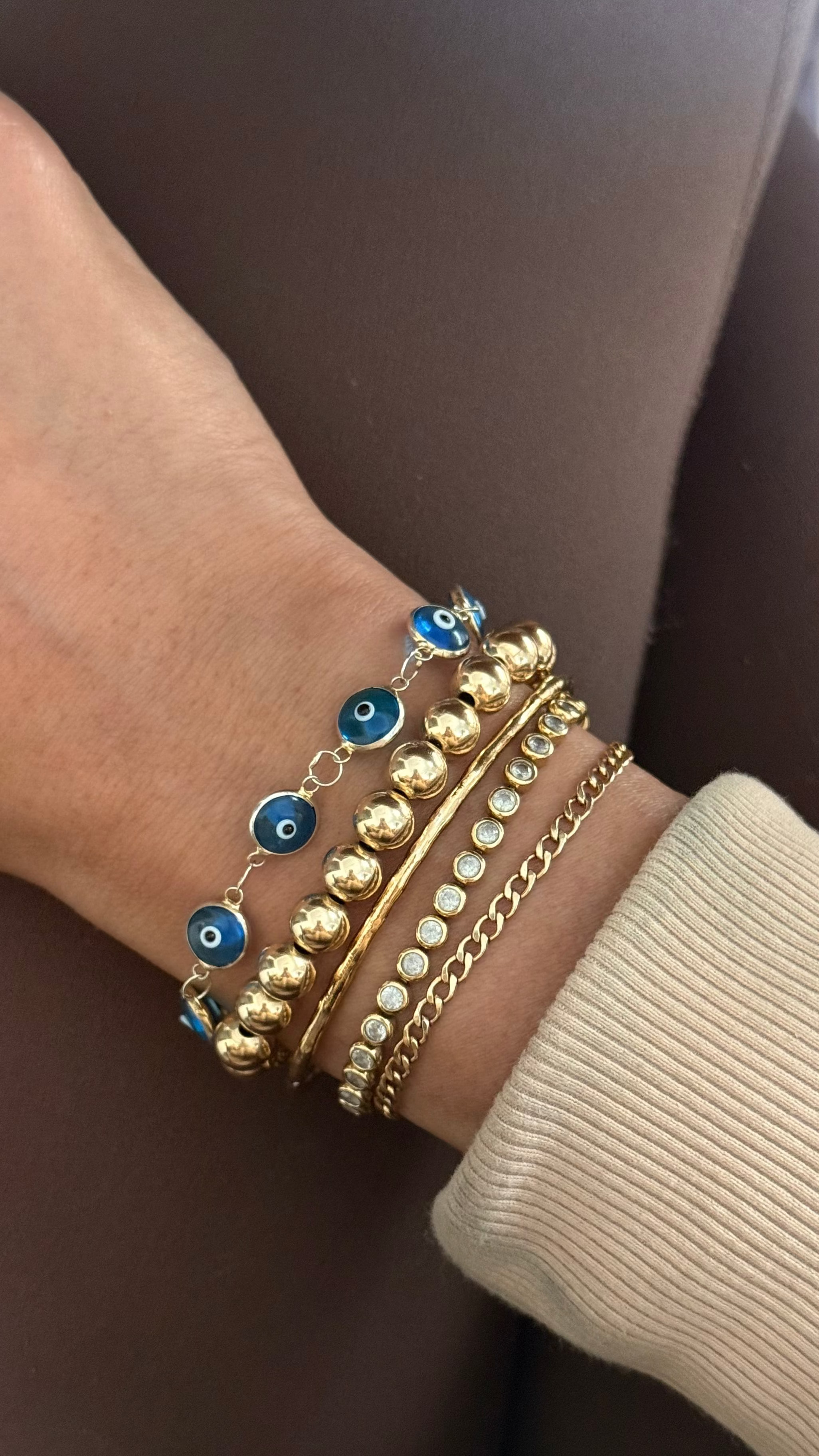Close up of a woman's wrist with a tan jacket and assorted gold bracelets on a brown background 