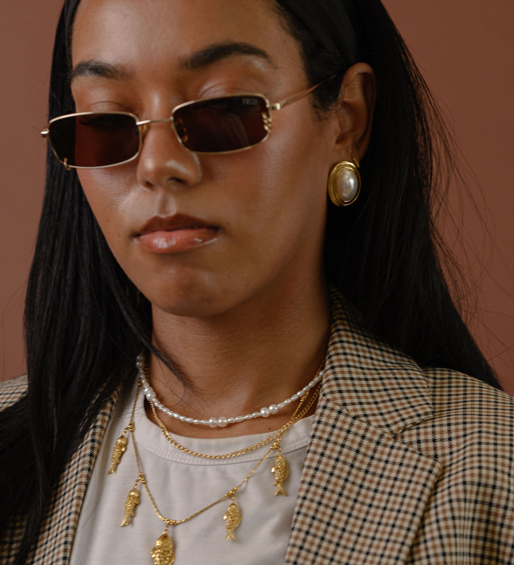 A woman in a blazer wearing statement gold earrings, sunglasses, and necklaces 
