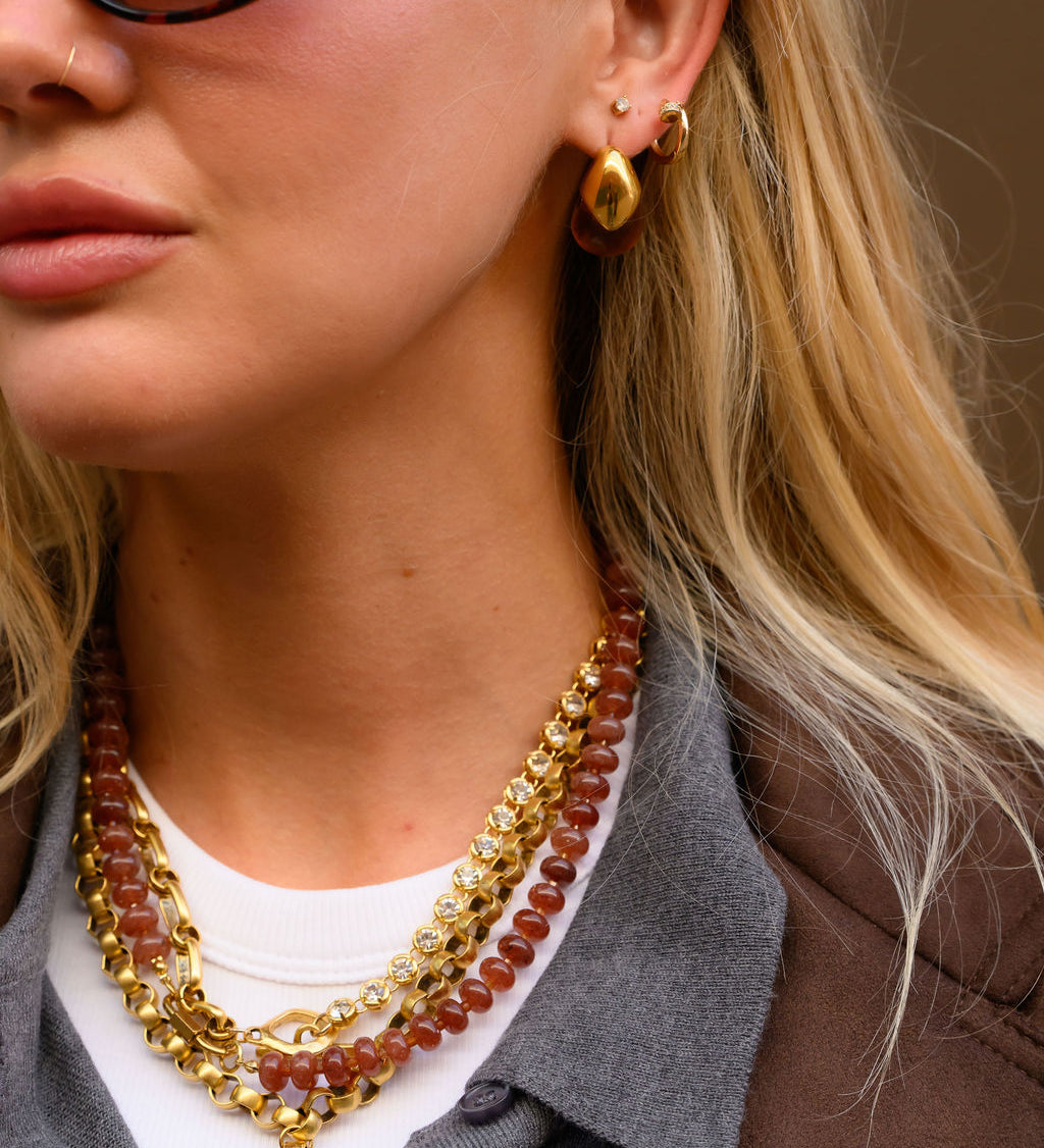 A woman wearing gold jewelry and sunglasses against a brown wall